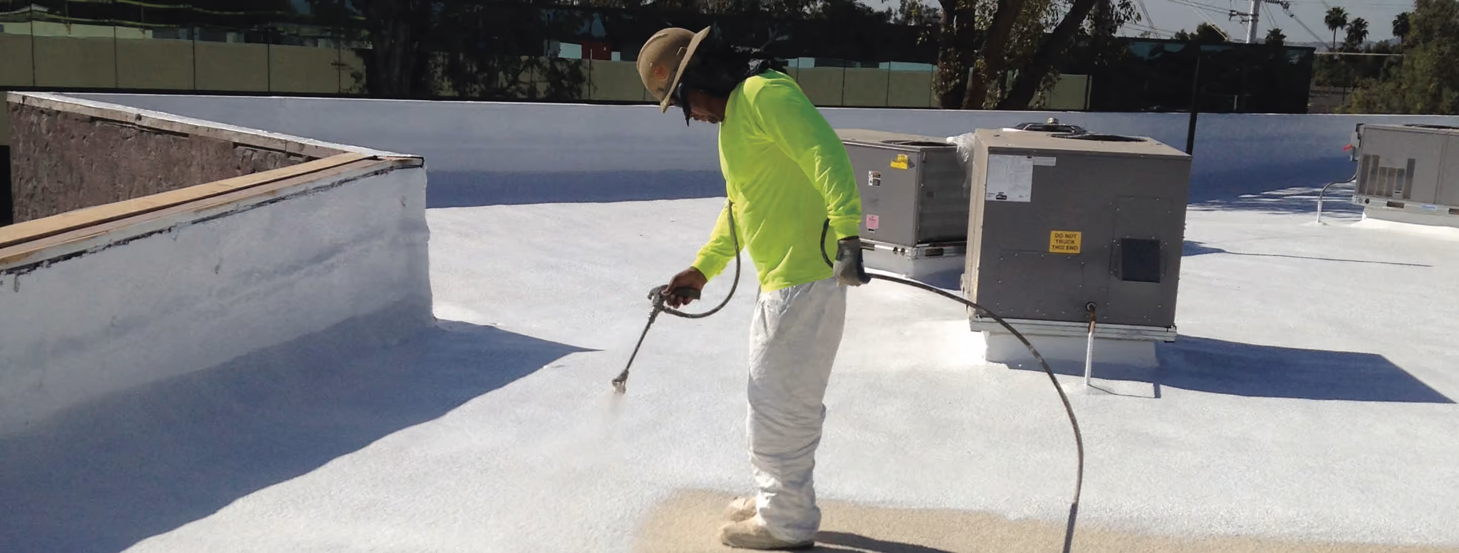 A worker spraying white coating across a flat roof, with a coated parapet wall on the left and rooftop air conditioning units on the right