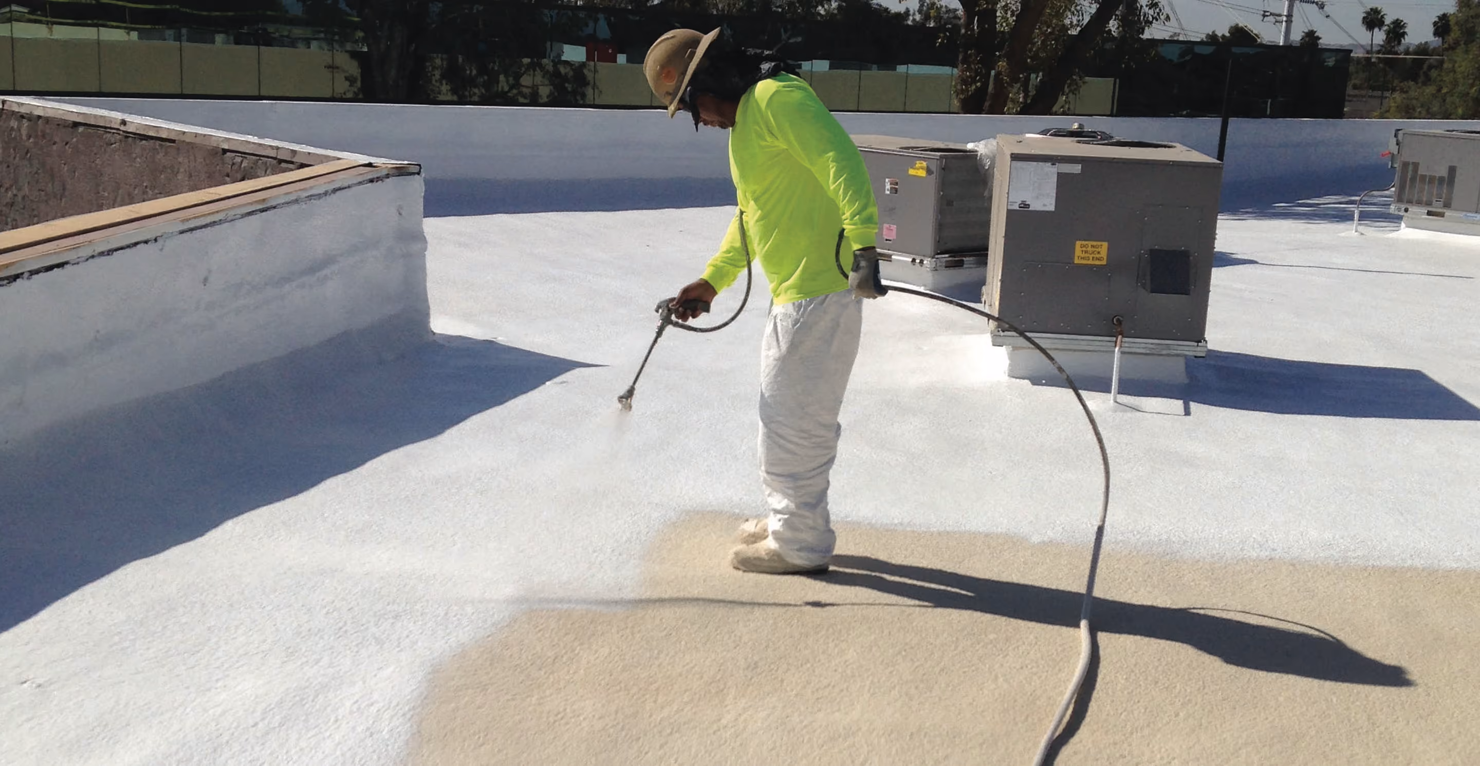 A worker in a bright yellow shirt spraying white coating over a flat roof, a coated parapet wall on the left and rooftop air conditioning units on the right