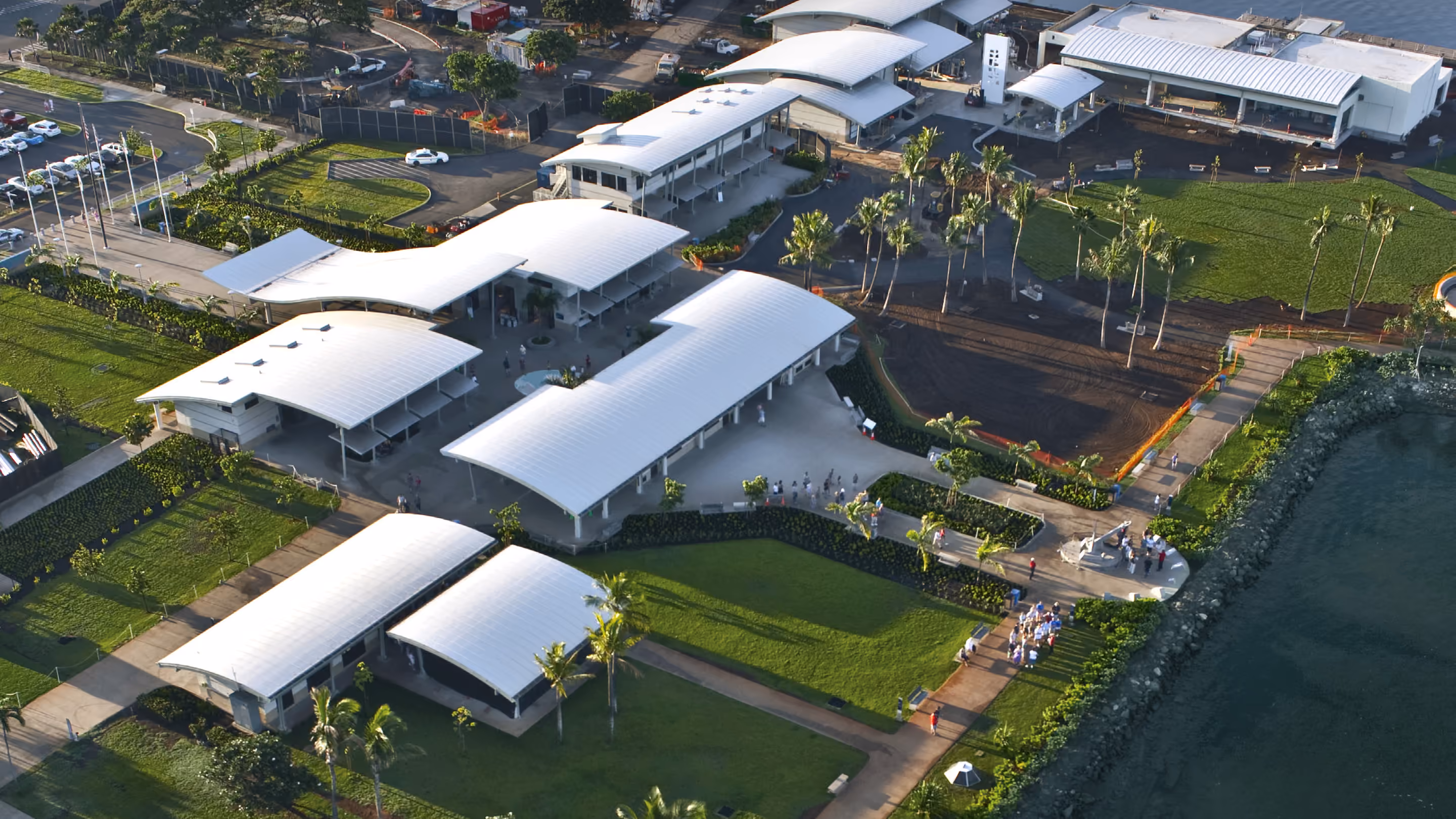 Low buildings under white curved roofs beside the water, seen from above, with palms, lawns, a parking lot and a walkway along the shore