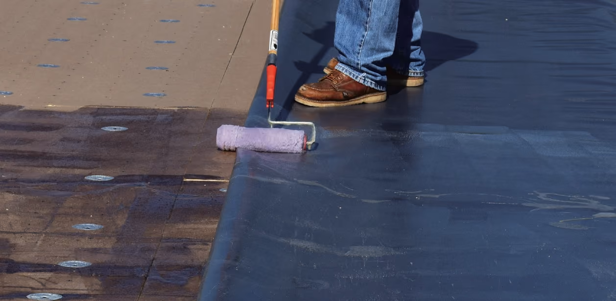 A roller spreading bonding adhesive across the roof deck beside fastener plates in the insulation board