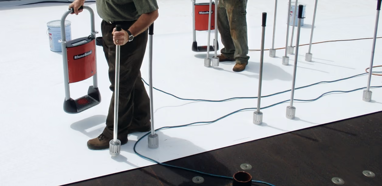 A crew induction-welding a white TPO sheet to the fastener plates under it, the welding machine on a hand cart and cooling magnets standing on the sheet