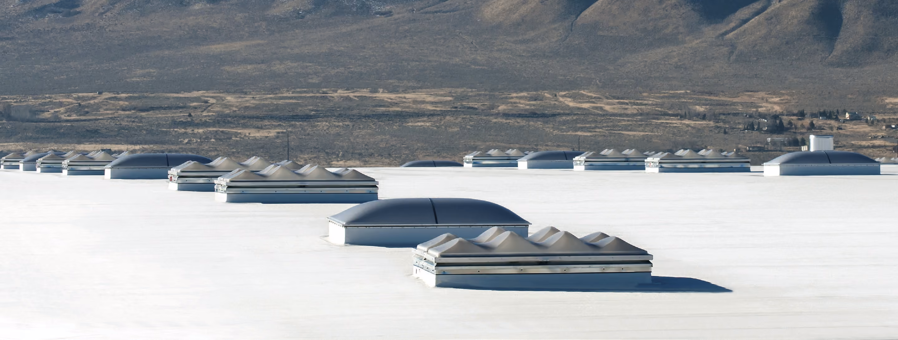 Rows of domed and ridged skylights on raised frames across a wide white flat roof, with a dry valley and mountains behind