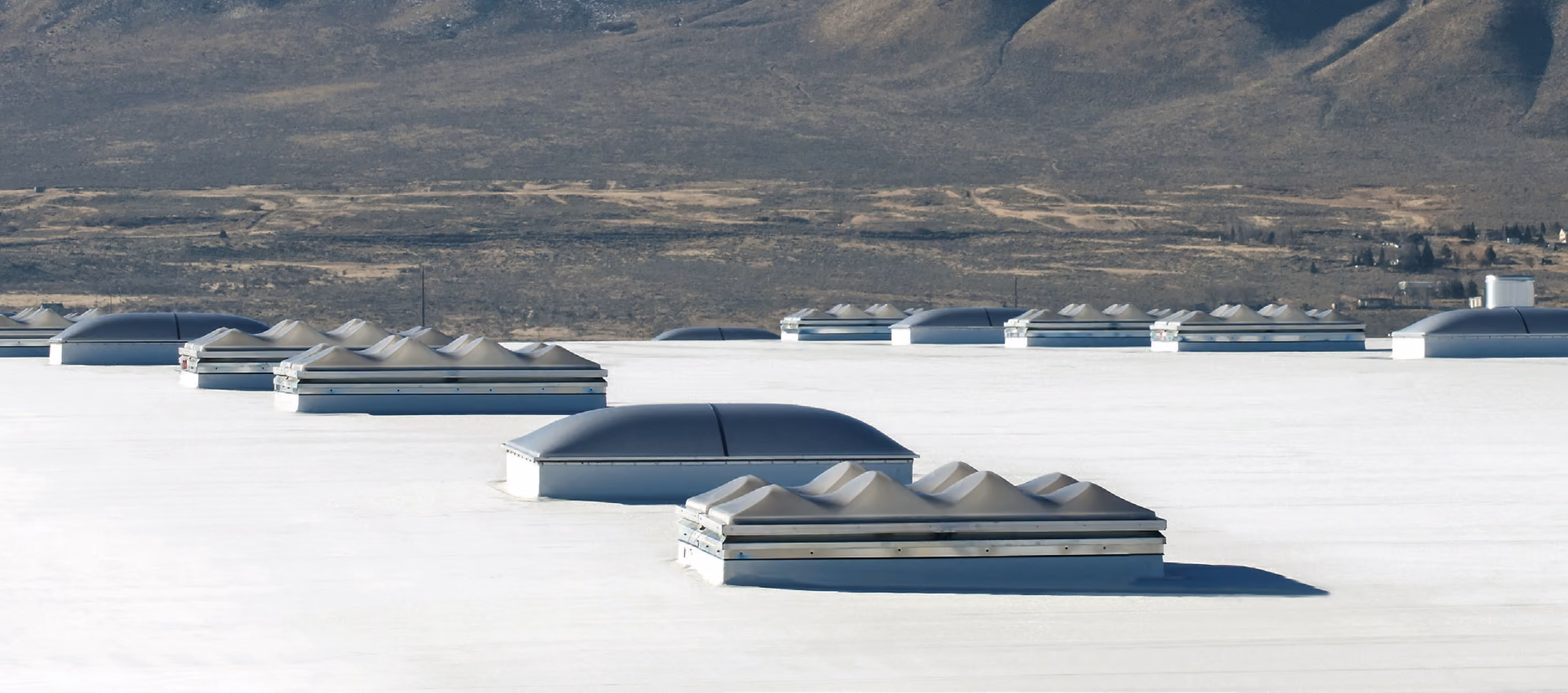 Domed and ridged skylights set in rows across a white flat roof, with a valley and mountains beyond