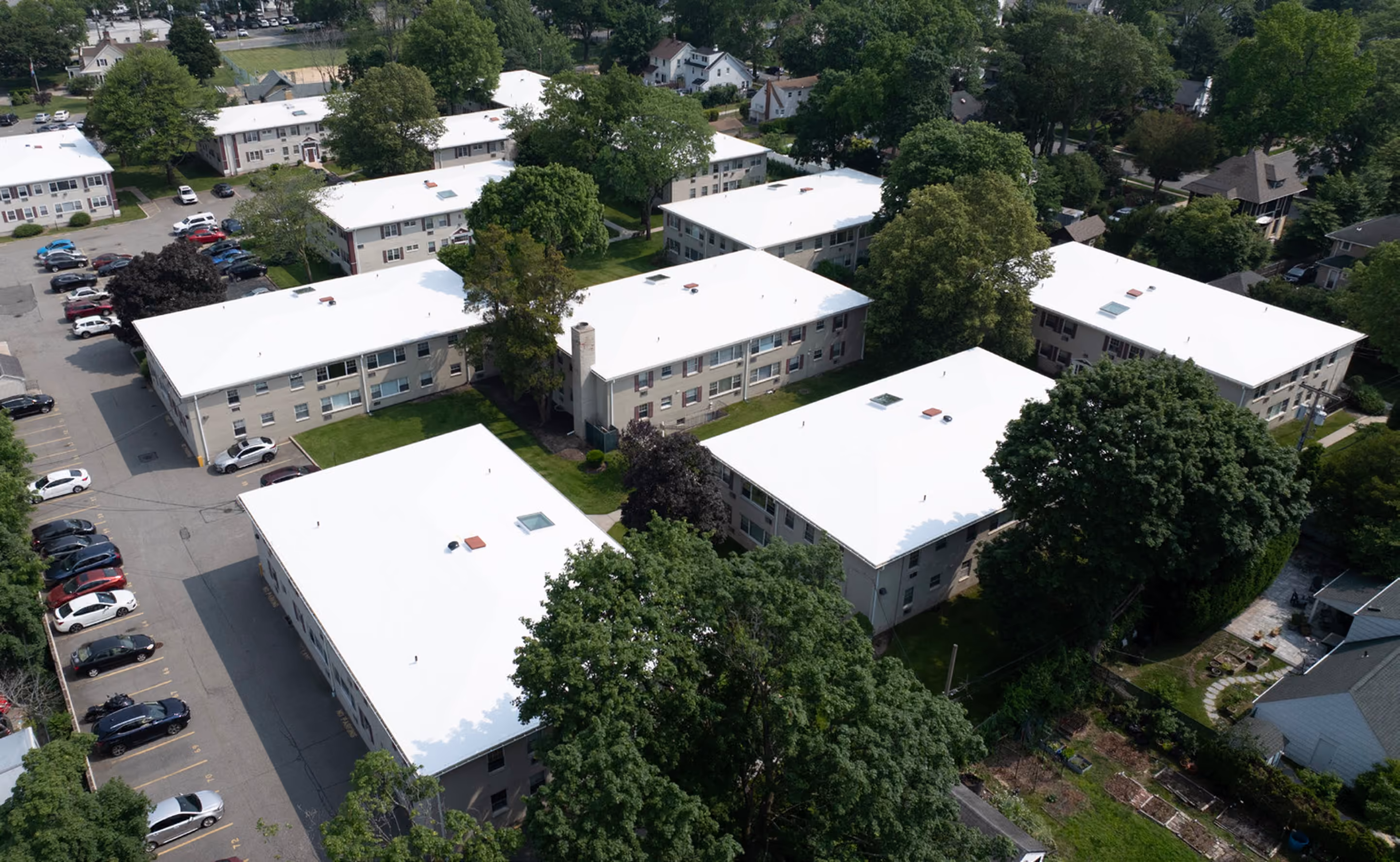 Two-story apartment buildings under new white TPO roofs seen from above, a parking lot along one side and mature trees between the buildings