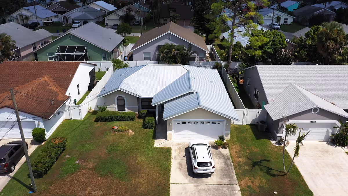 A white standing seam metal roof over a single-story house, with a car in the drive and neighboring homes along the street