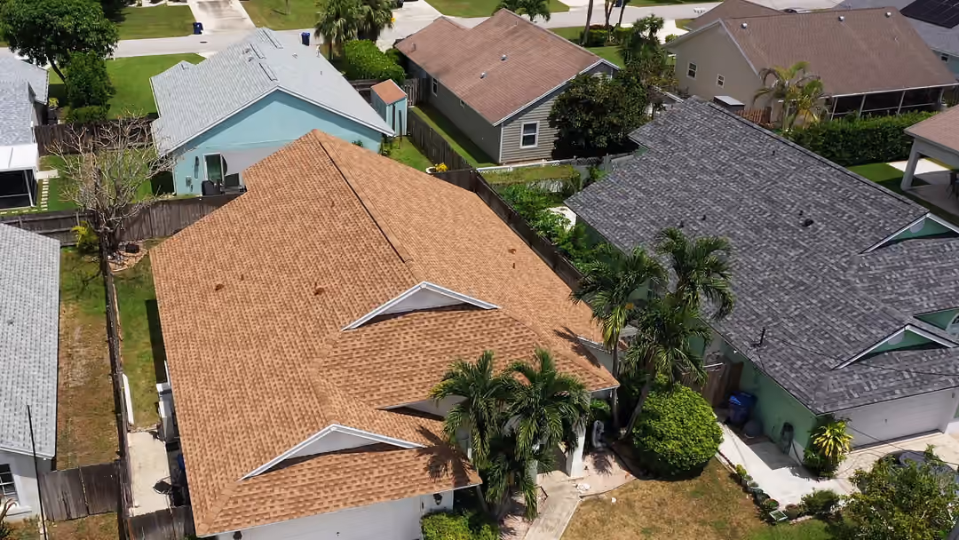 A tan shingle roof over a single-story house, hipped with two front gables and royal palms standing along the entry walk