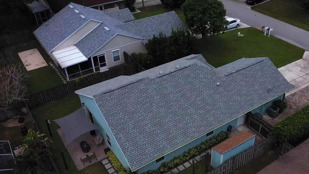 A long gray shingle roof over a single-story mint-green house, with a shade sail stretched over the paver patio at the side