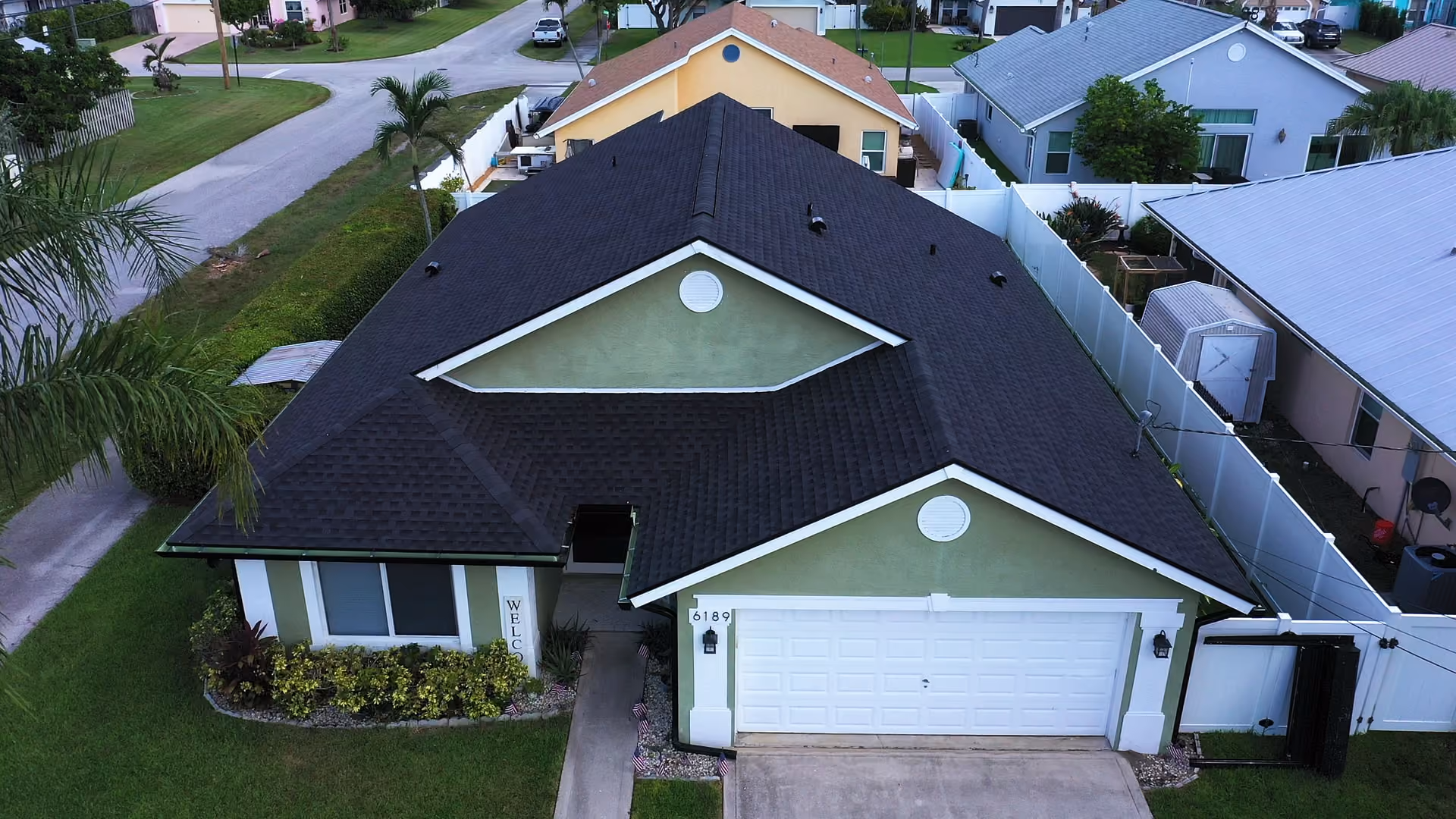 A sage-green single-story house under a new charcoal architectural shingle roof, with white gable trim and a low planting bed along the front