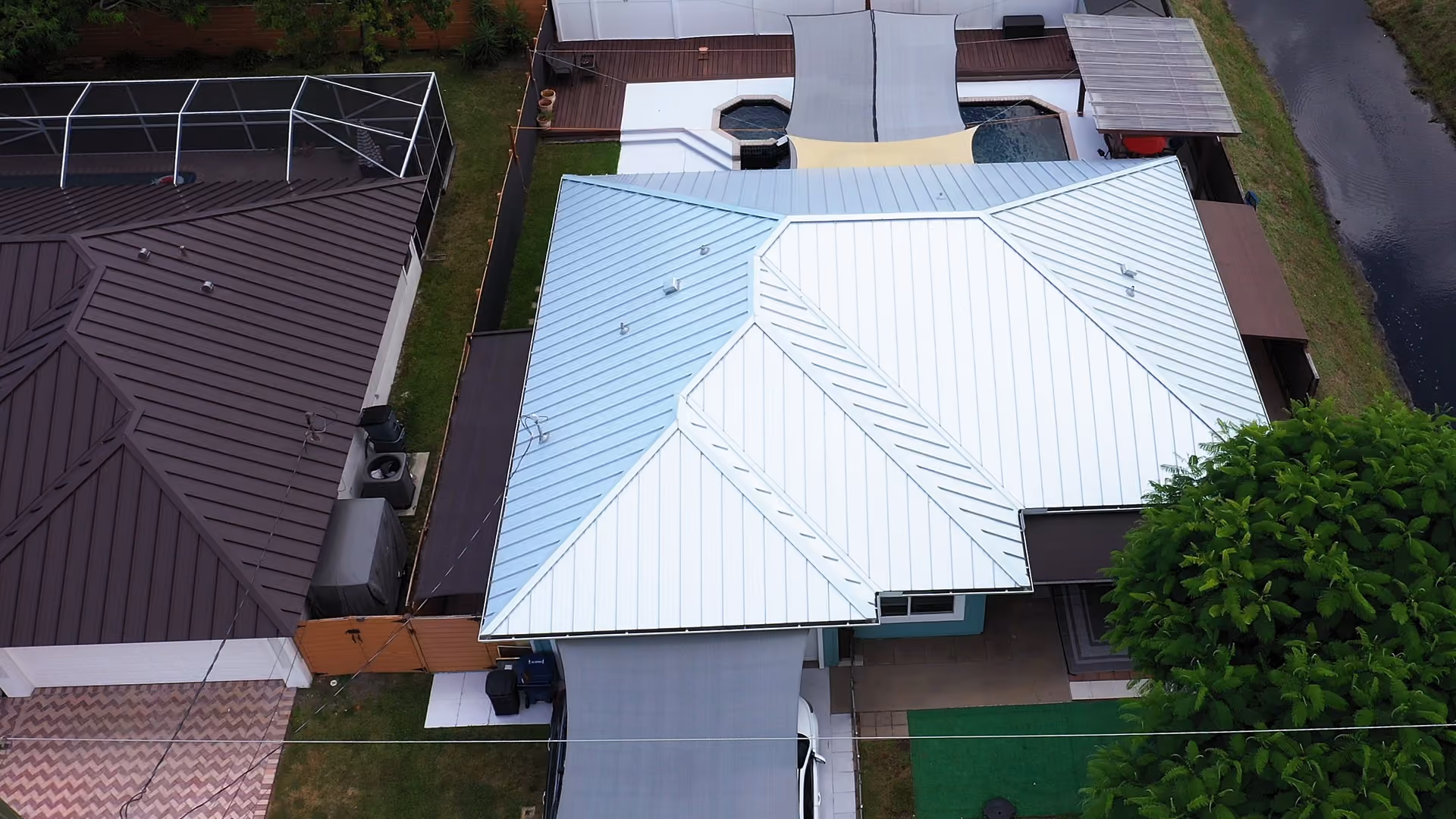 A pale silver standing seam metal roof seen from directly above, a canal running along the edge of the lot and a shaded pool deck behind the house