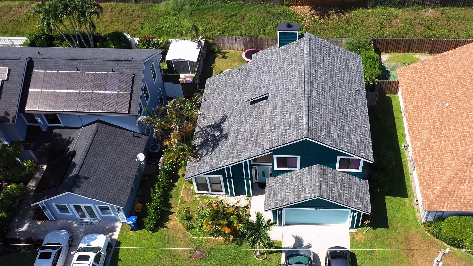 A two-story teal house with a gray blended architectural shingle roof and white trimmed gables, palms crowding the front walk