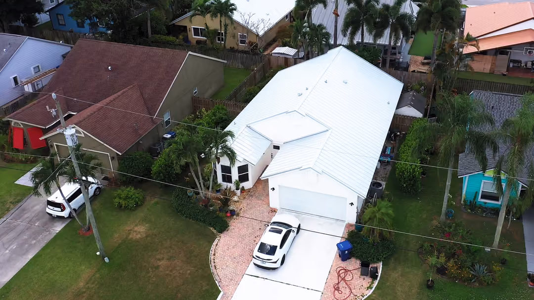 A white metal hip roof seen from above, with a brick paver driveway curving to the garage and royal palms along the lot line