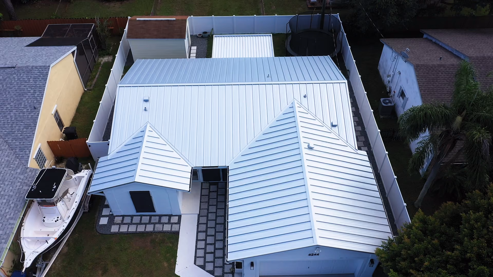 A white standing seam metal roof photographed straight down, with a center-console boat on a trailer parked along the fence at one side