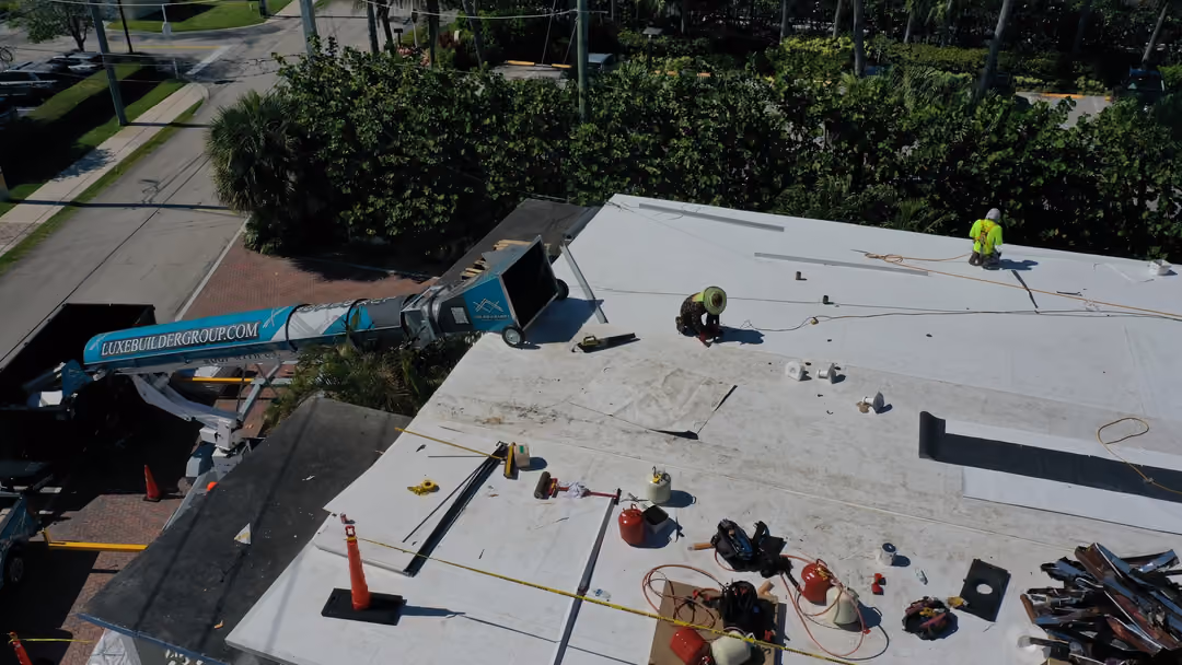 A material chute running from the truck up onto a white flat roof, with two roofers working at the top of the run