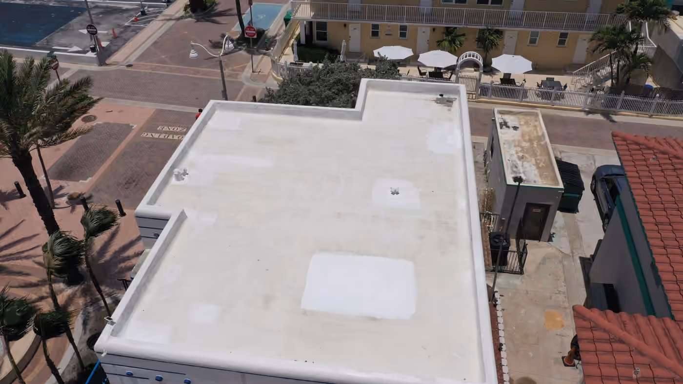 A small one-story corner building with a white flat roof inside a low parapet seen from above, with a brick-paved street corner in front of it