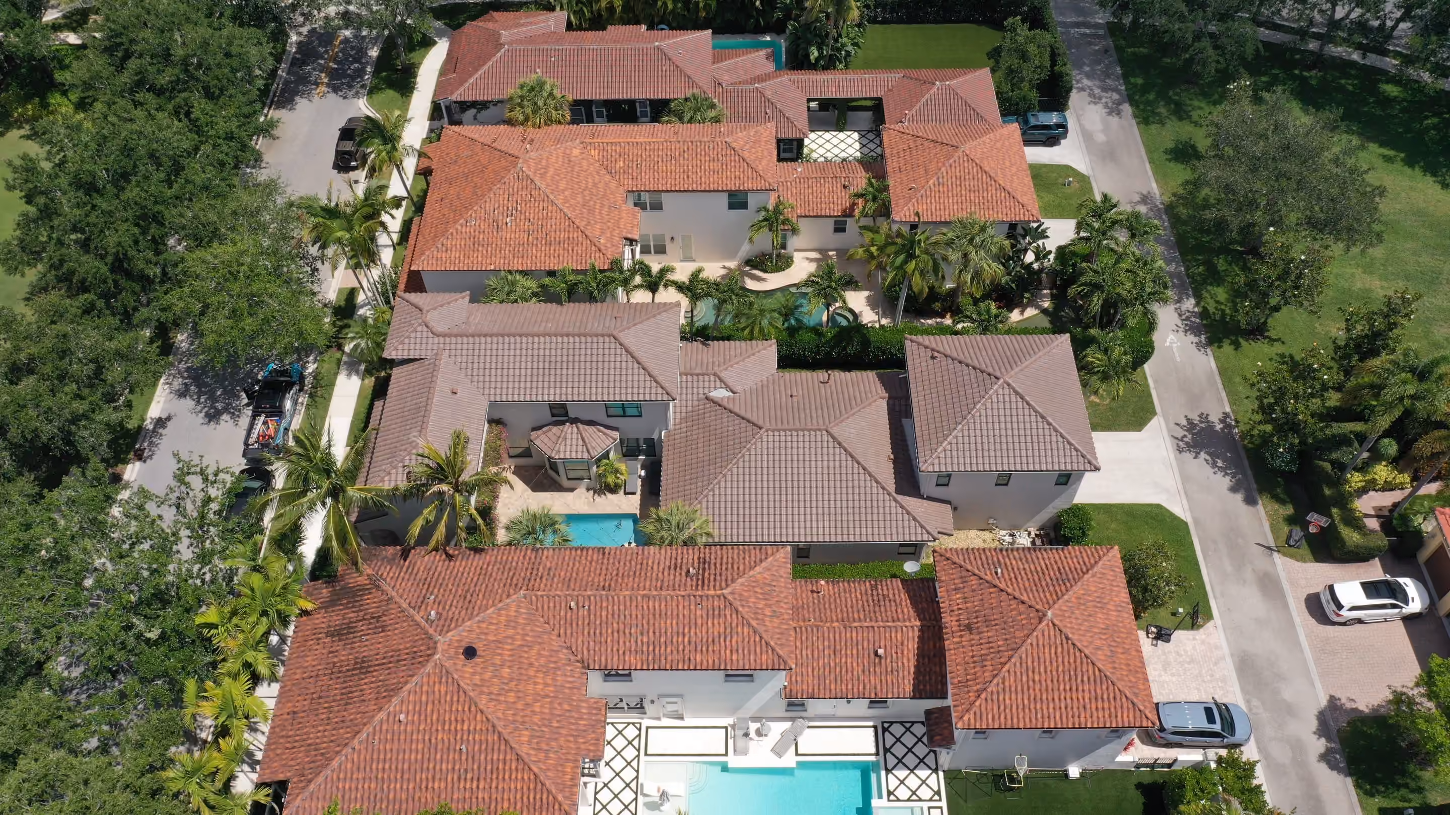 A run of neighboring homes with barrel tile roofs seen from above, screened pools between them and a paver street along one side