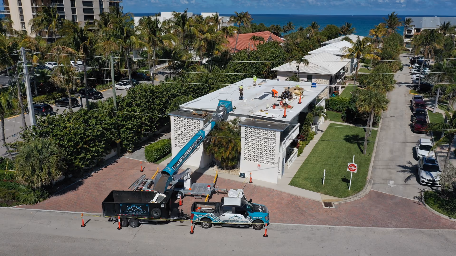 A beachside condo building with a white flat roof being replaced, the Luxe conveyor boom raised from a branded truck and trailer at the curb and crew working on the deck