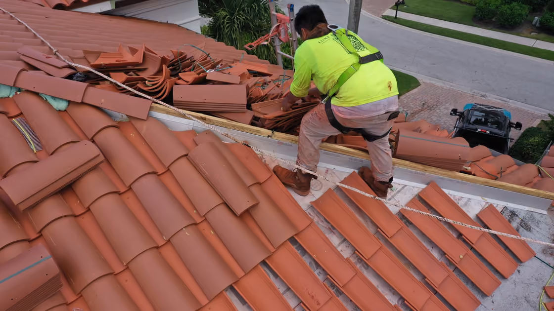 A roofer setting a course of clay barrel tile by hand, with stacked tile and battens across the roof beside him
