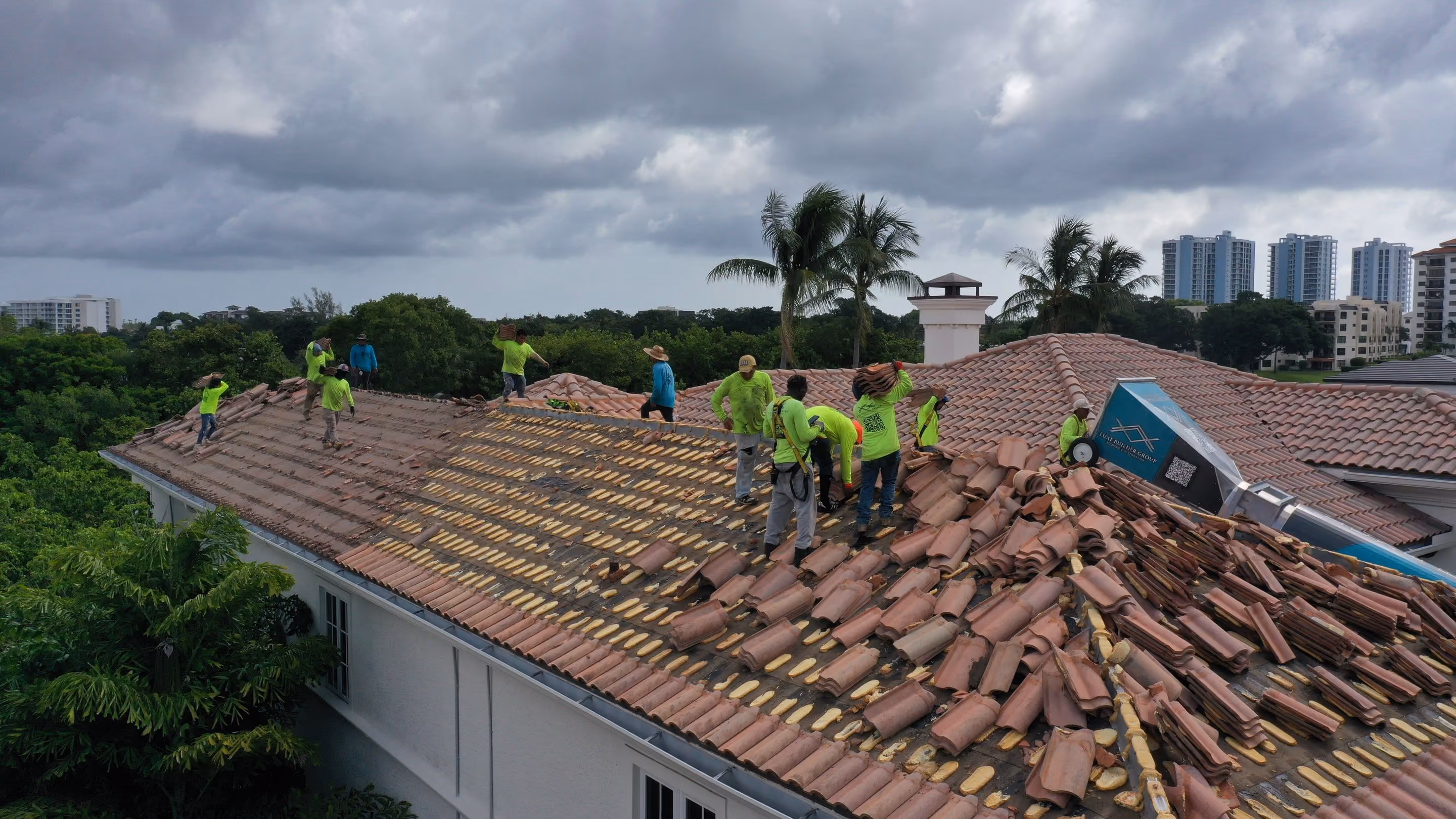 A crew of roofers working across a large barrel-tile roof, lifting the old tile into stacks beside a Luxe debris chute under an overcast sky