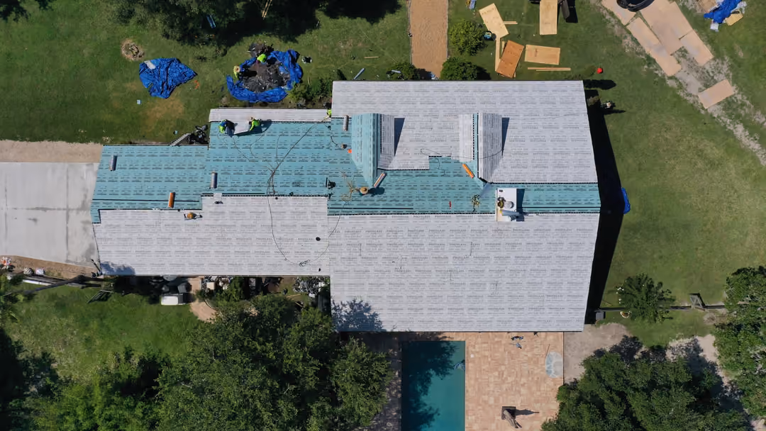 A large roof seen from directly above after dry-in, gray underlayment with sealed green seams covering every plane and a crew working at one corner