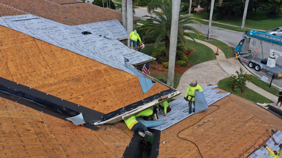 Crew members laying underlayment over a bare plywood roof deck, with a Luxe-branded trailer parked in the driveway below
