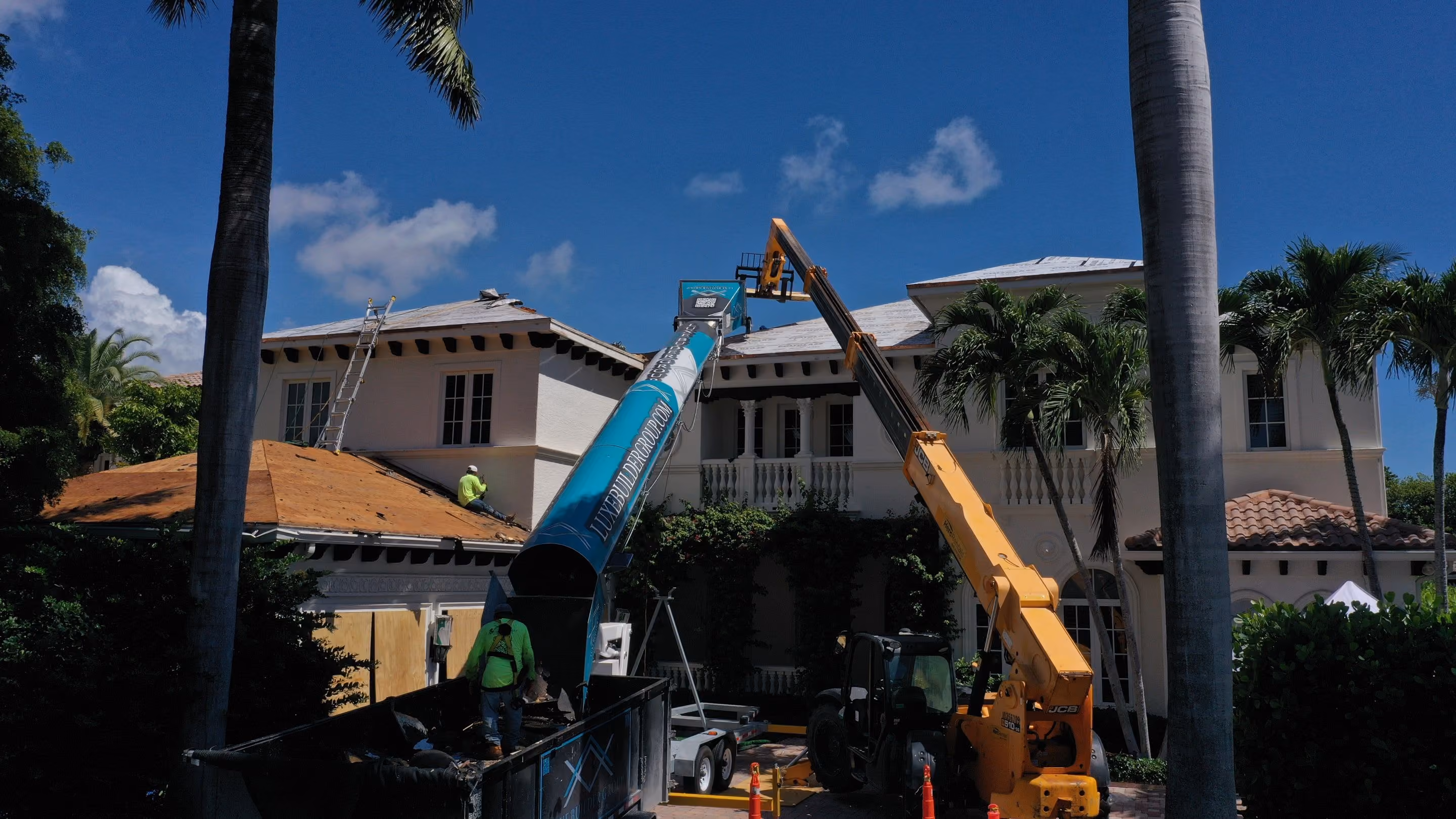 A two-story white estate with a columned loggia and a balcony balustrade, its roof stripped to bare decking, with a telehandler and a material conveyor set up in the front drive