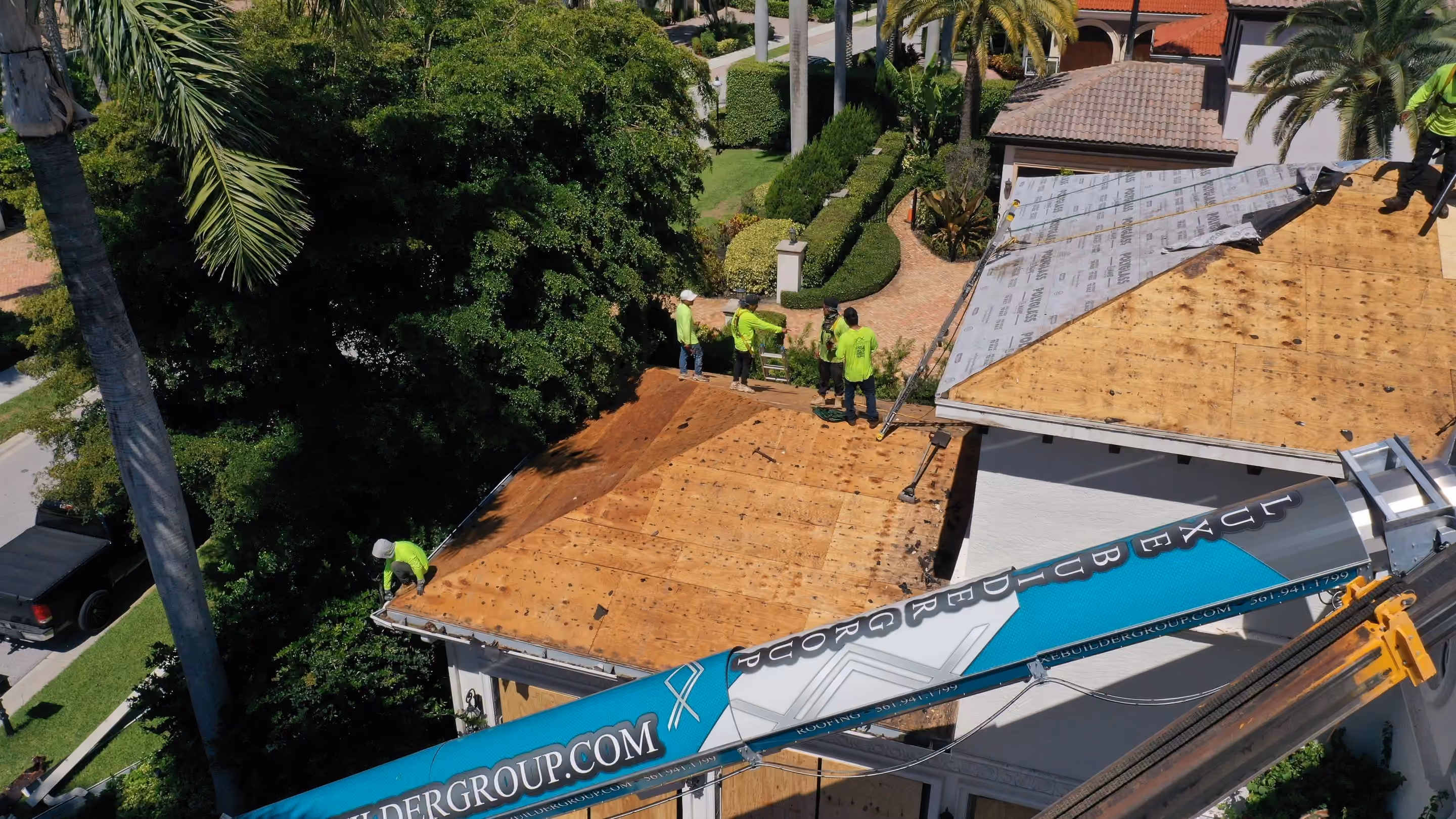 A crew standing on an estate roof stripped to bare decking, with a Luxe Builder Group boom conveyor raised to the eave from the drive below
