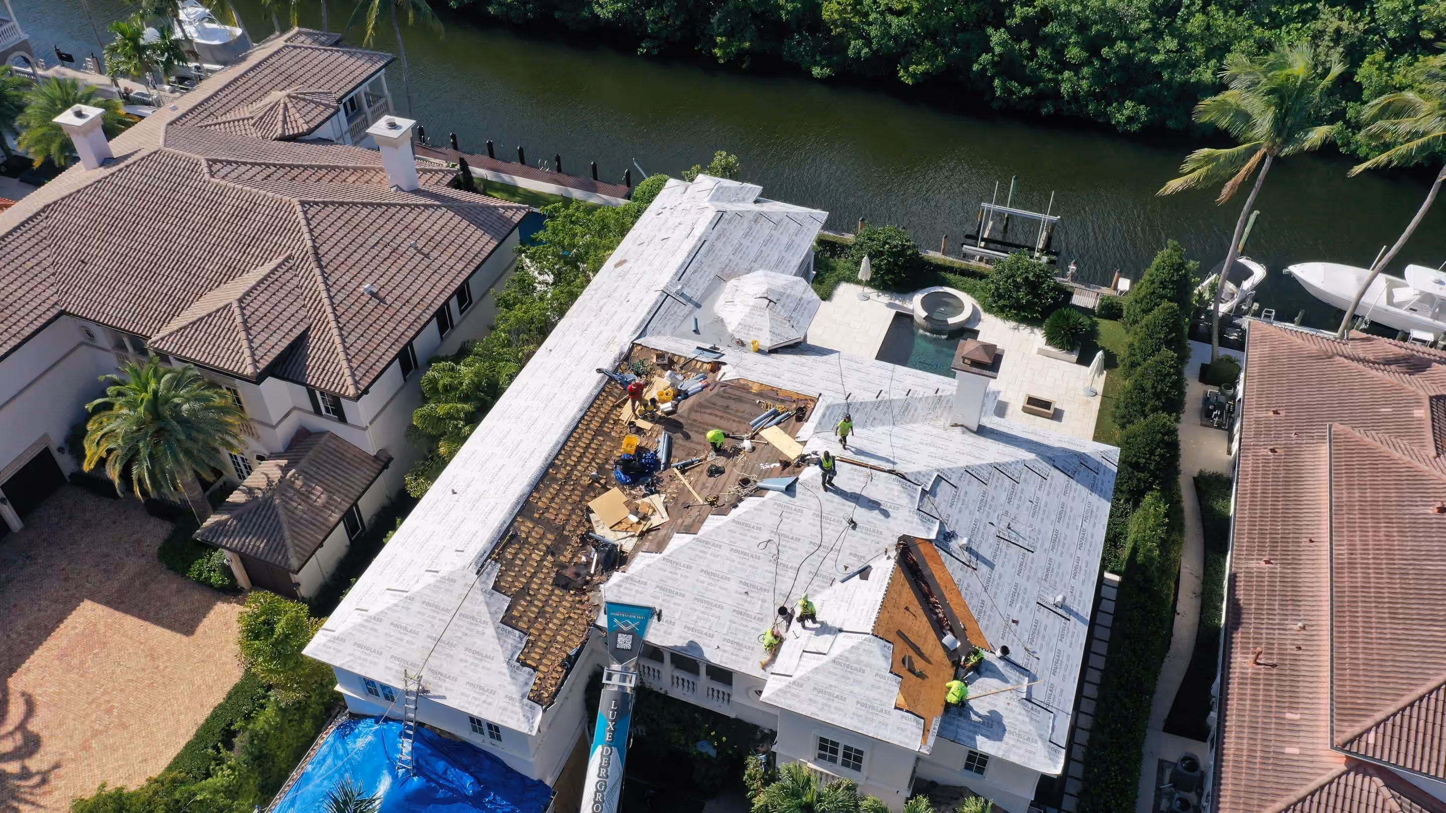 A waterfront estate mid-replacement with the crew working across the roof, a private dock and a moored boat on the canal behind