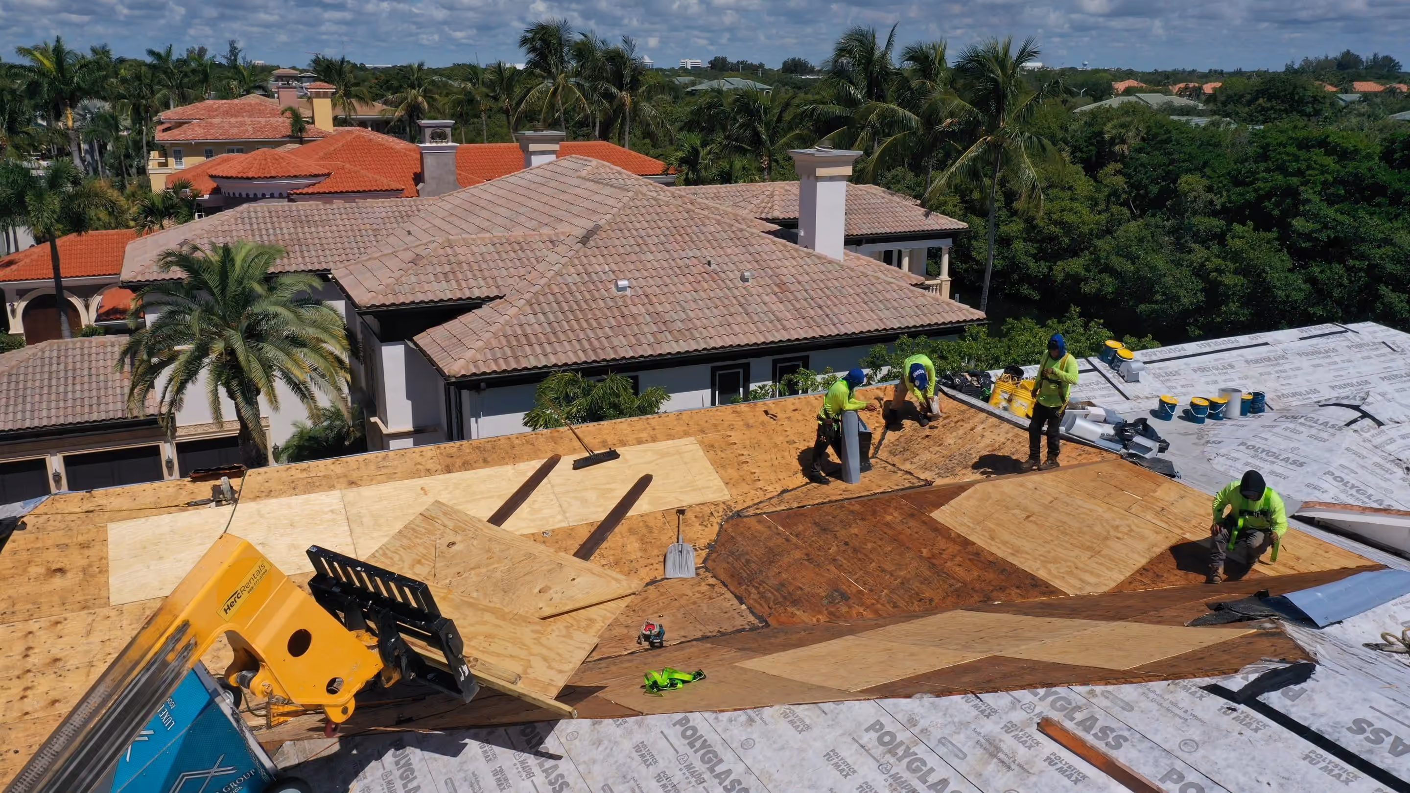 A crew working across an estate roof stripped to plywood and underlayment, with a second large tile-roofed house directly behind it