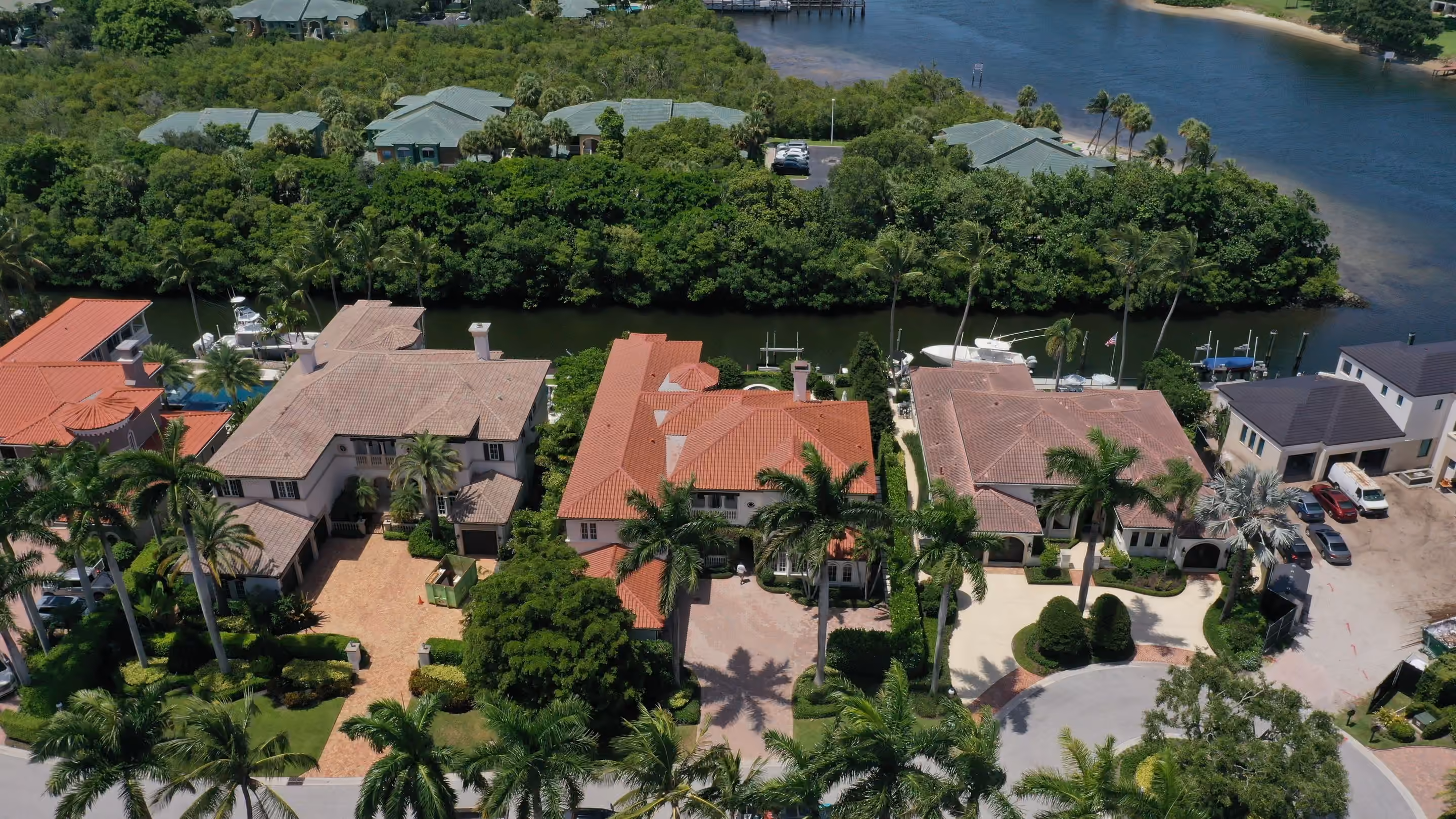 Three large waterfront homes side by side under clay barrel-tile roofs, the middle roof newly laid, with a mangrove channel and docked boats behind them
