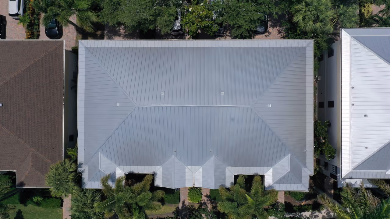 A silver-gray metal roof with raised seams seen from directly above, hipped at both ends, with a row of small dormers along one edge and palms in front of the building