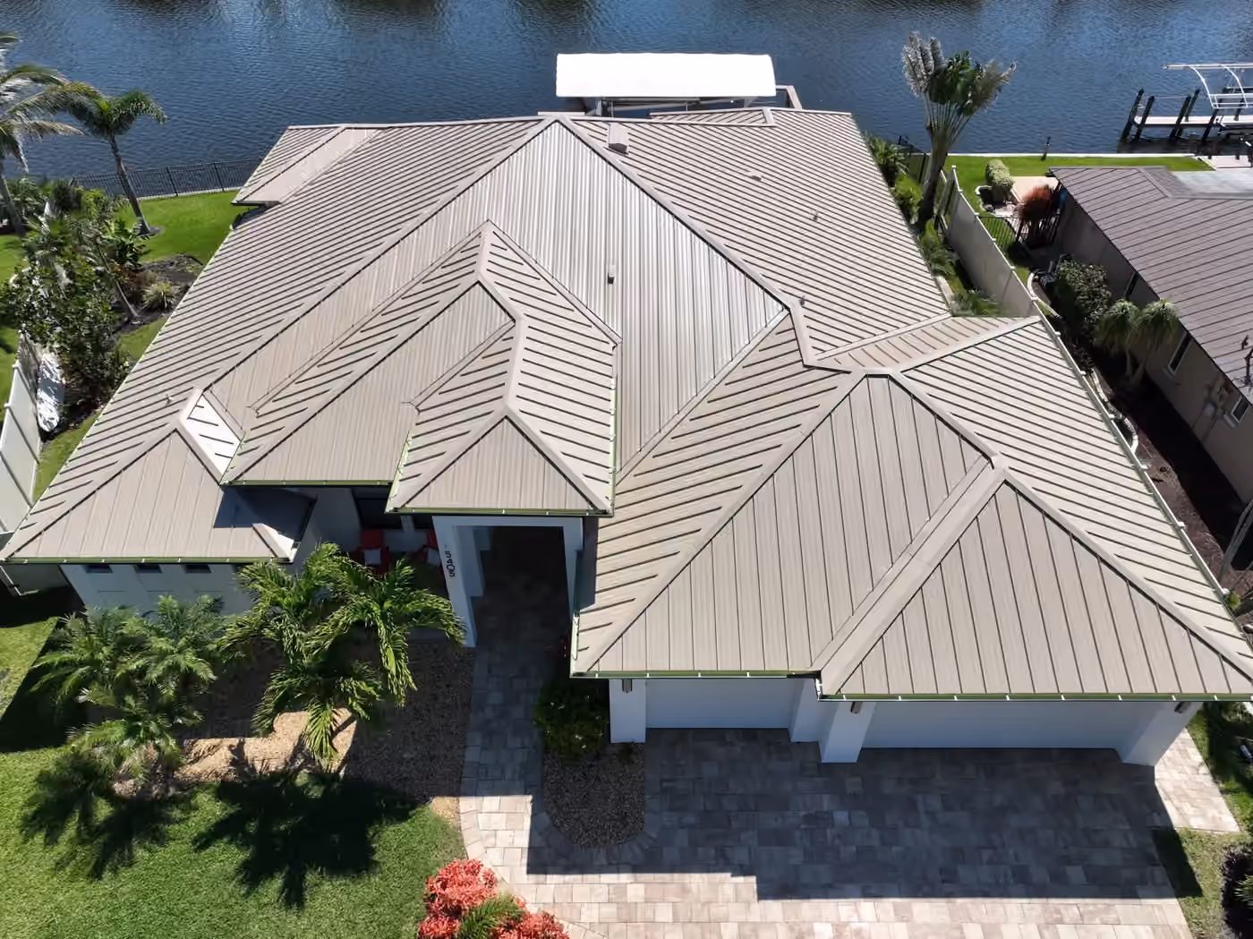 A large white waterfront house under a new taupe standing seam metal roof, with a paver drive in front and a covered boat lift on the canal behind it