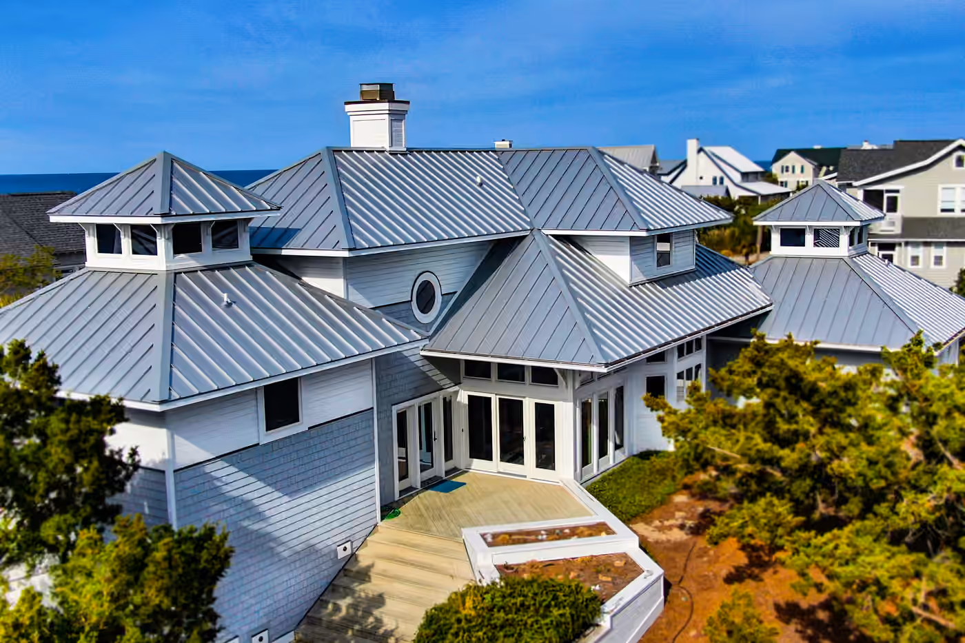 A large gray-blue coastal house under a new silver standing seam metal roof, with two white cupolas rising from the hips and the ocean beyond