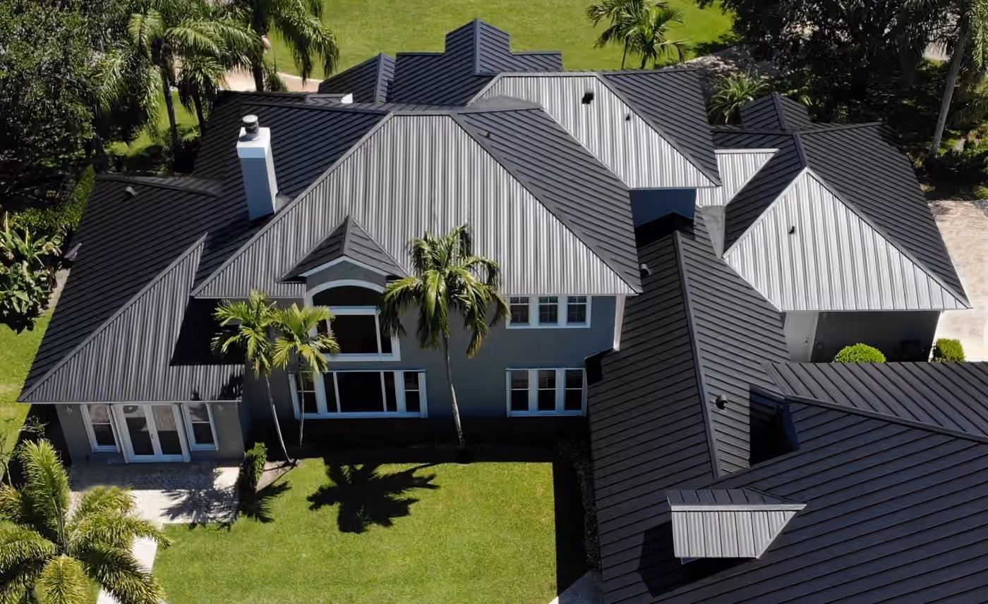 A large two-story gray house under a new charcoal standing seam metal roof, with gabled wings, a white chimney and palms across the lawn