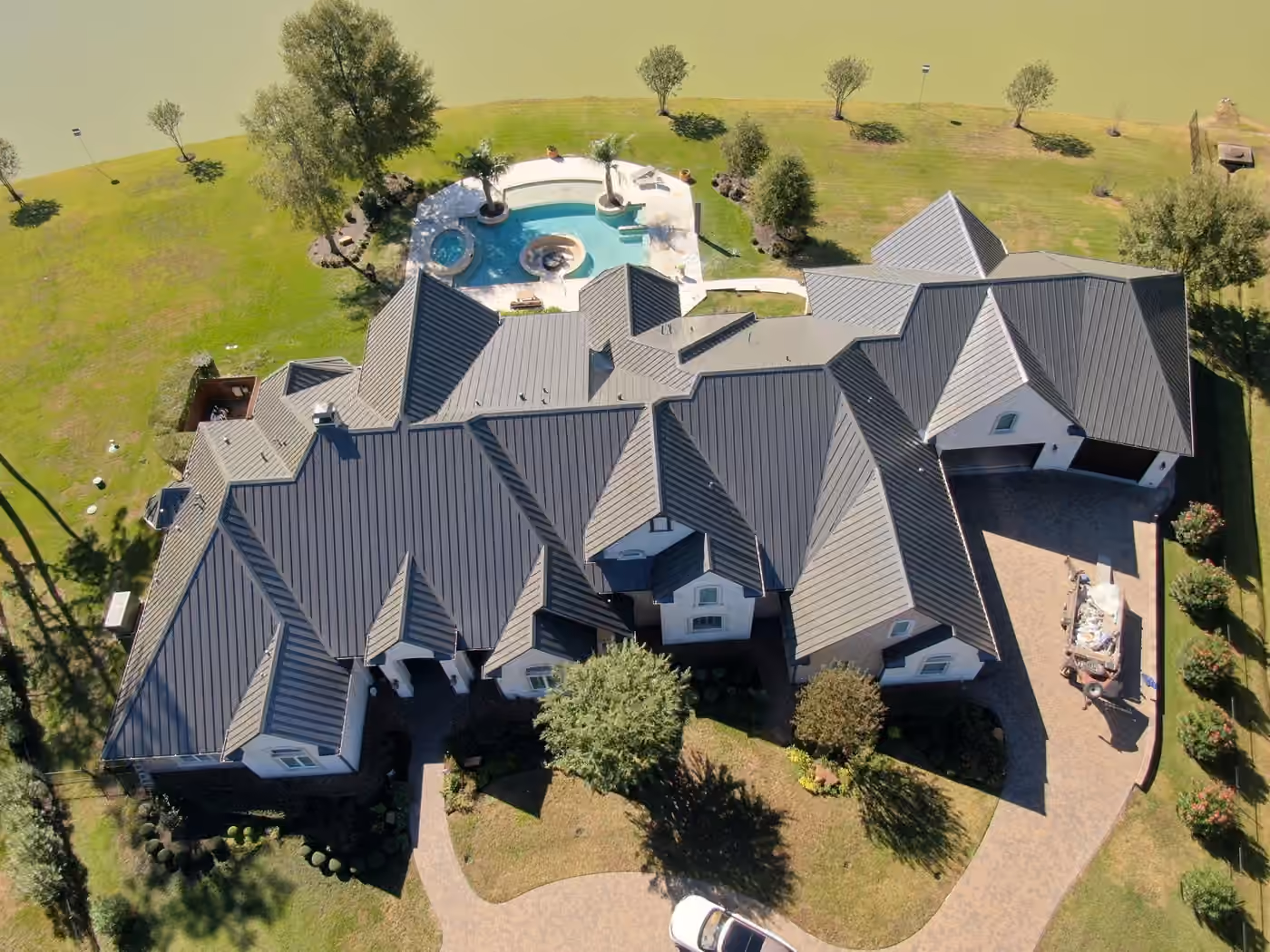 A large stone and stucco estate under a new charcoal standing seam metal roof seen from above, with a free-form pool and spa on the lawn running down to the lake