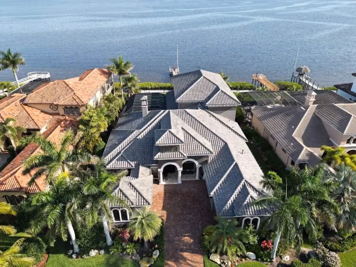 A large waterfront estate under a new gray flat-tile roof seen from above, with an arched entry loggia, a paver drive between royal palms, and boat docks on the bay behind it