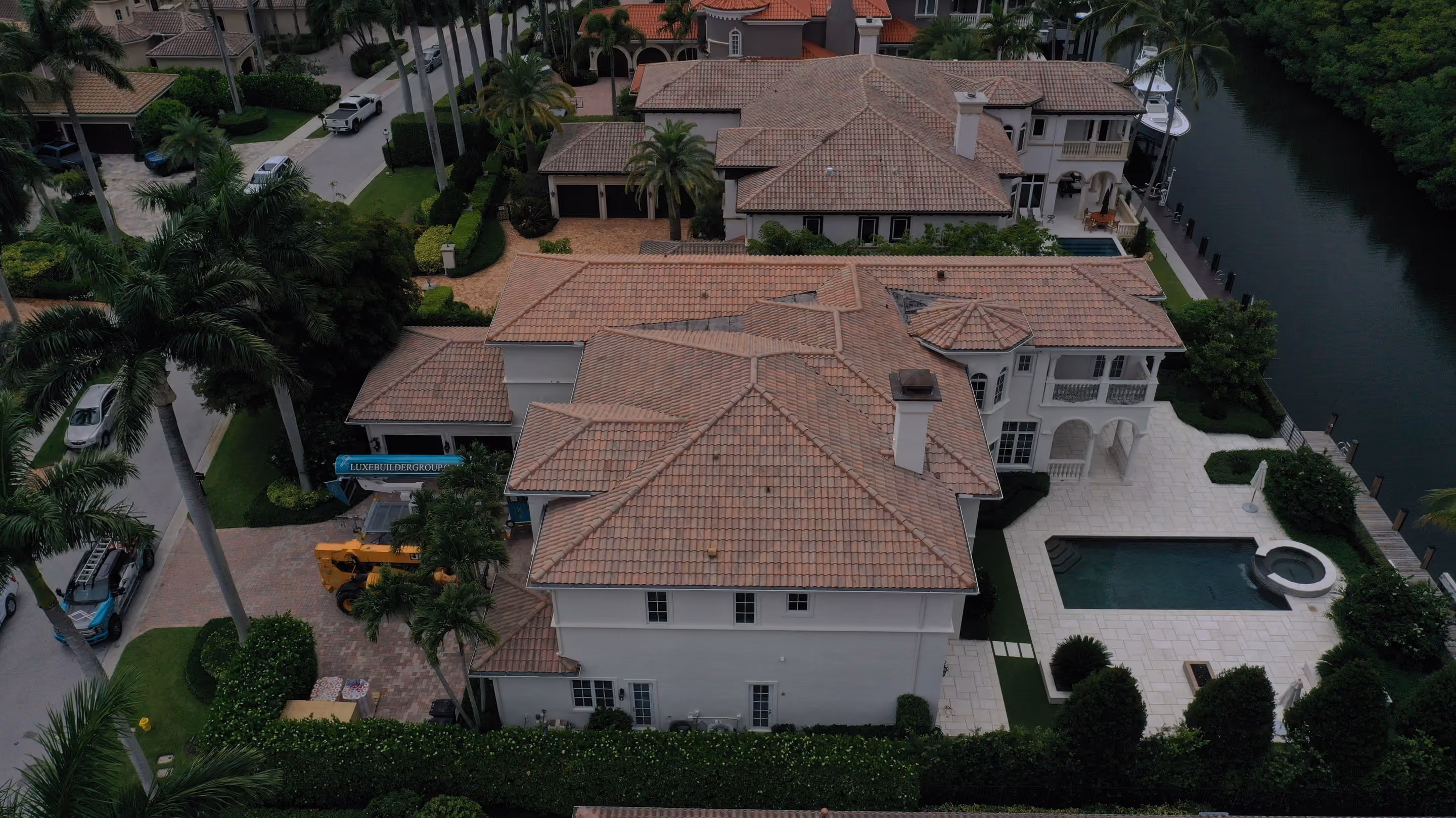 A two-story home under a finished clay tile roof beside a canal, with a pool and raised spa on the stone deck and a Luxe chute and loader on the paver drive