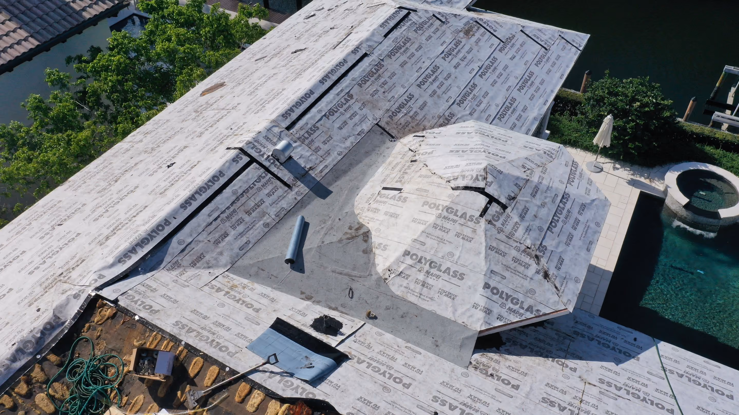 An estate roof stripped back to underlayment across every slope, with the pool and spa deck at the edge of the frame
