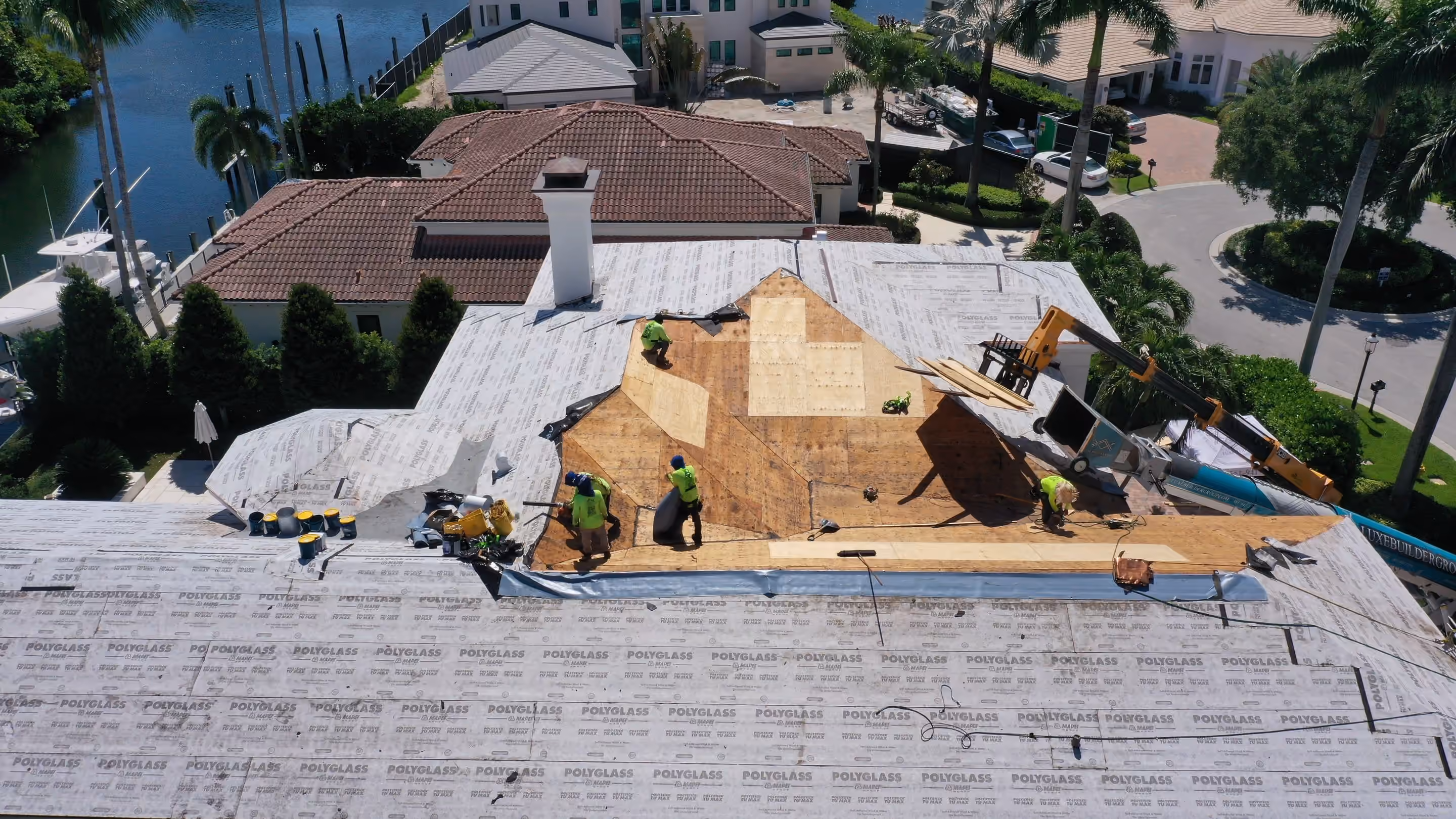 A crew working on a large estate roof opened to bare plywood, with the surrounding slopes already covered in underlayment and a waterway behind the house