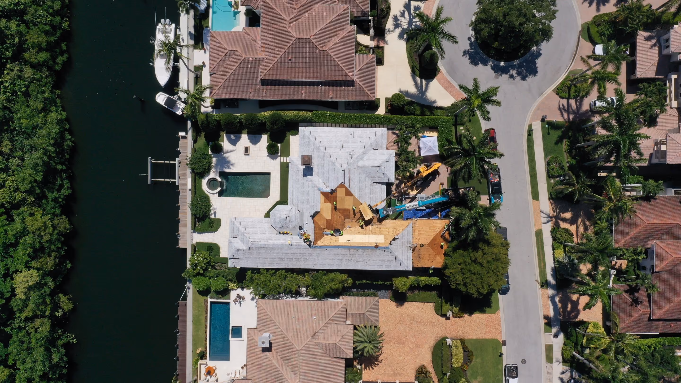 A large waterfront home seen from directly above during a roof replacement, most of the deck covered in underlayment with one section stripped to bare plywood and roofers working across it