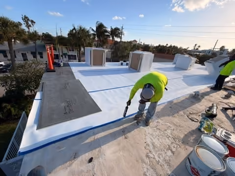 Roofers rolling out TPO membrane across a flat roof deck, the cover board still exposed ahead of them