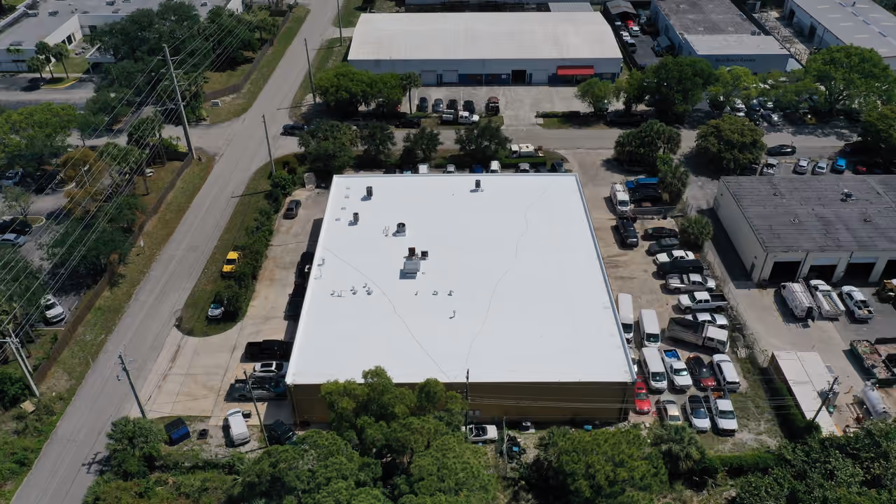 A warehouse with a white flat roof seen from high behind it, with service lots full of trucks and vans around it and other industrial buildings beyond