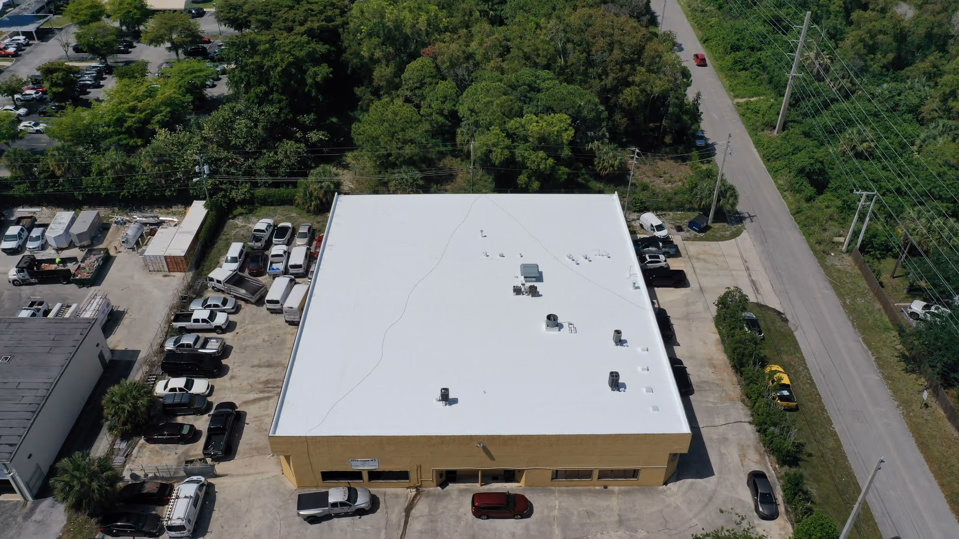 A tan warehouse with a white flat roof seen from the air, with pickups, vans and cars parked around it, a road and power lines along one side and thick trees behind
