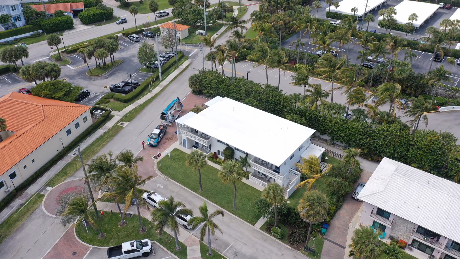 A two-story white building with a white flat roof and balconies, seen from the air on a corner lot with palms, a Luxe Builder Group truck and a trailer carrying a teal chute parked on the brick drive beside it
