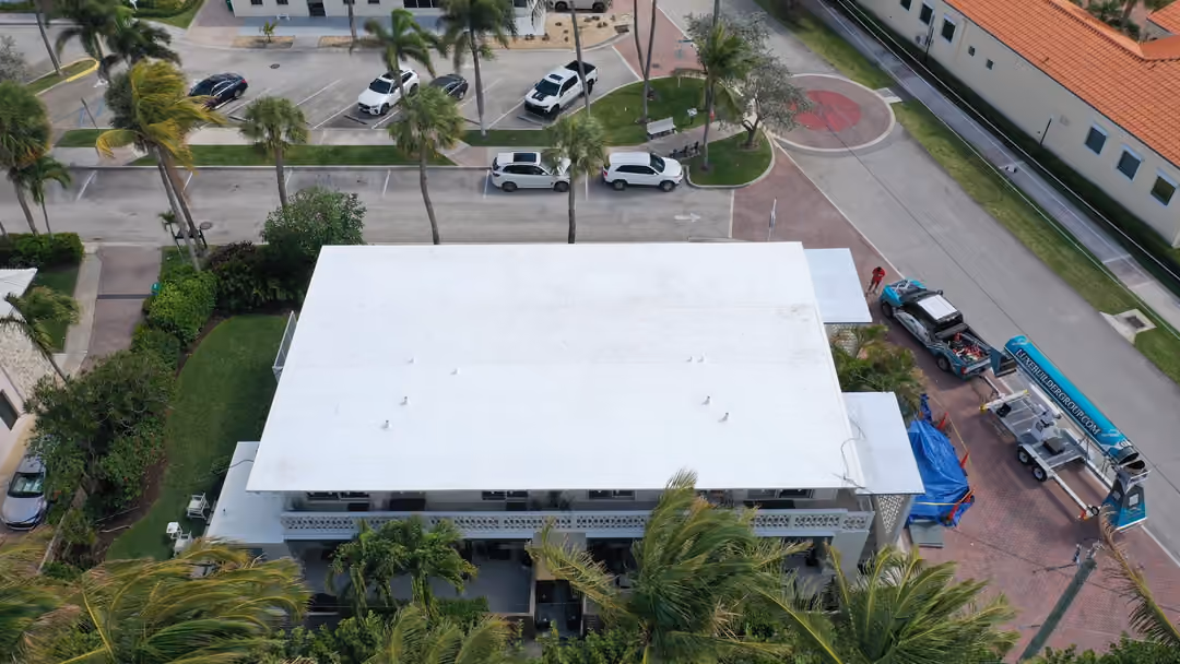 A white flat roof on a two-story condo building in a managed community, parking bays and palms around it, a Luxe truck and conveyor staged on the drive alongside