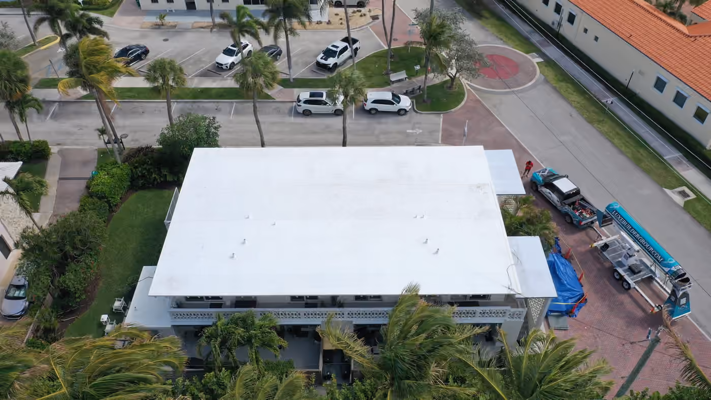 A white flat roof on a two-story multi-unit building seen from above the front, with balconies below it, palms around it and a truck and trailer on the brick drive beside it