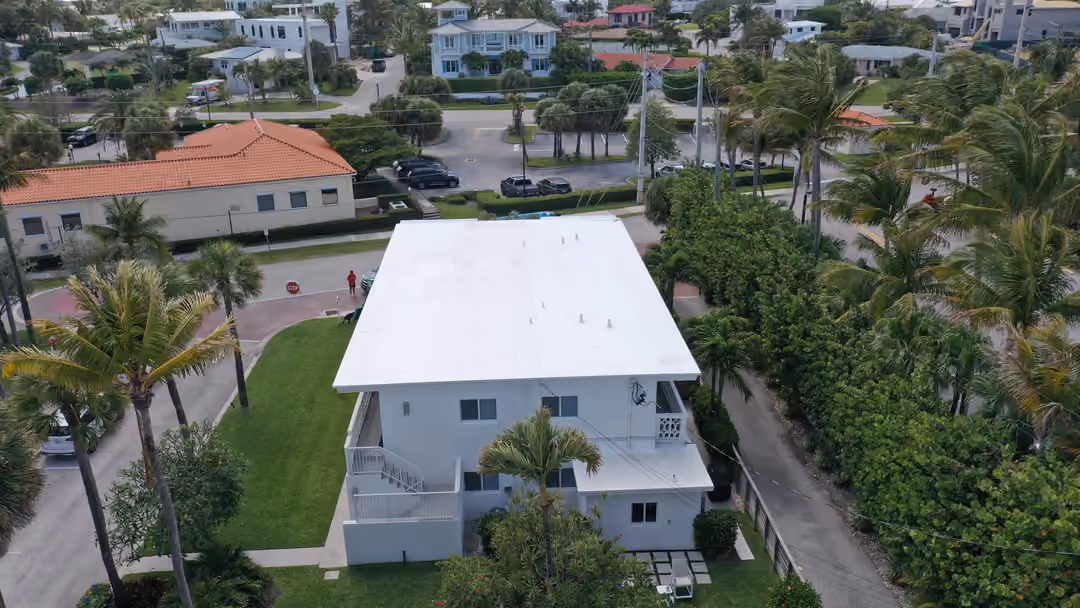 A white flat roof on a two-story condo building seen from above, vent pipes standing up across the field, an exterior stair down one side and the street corner beside it