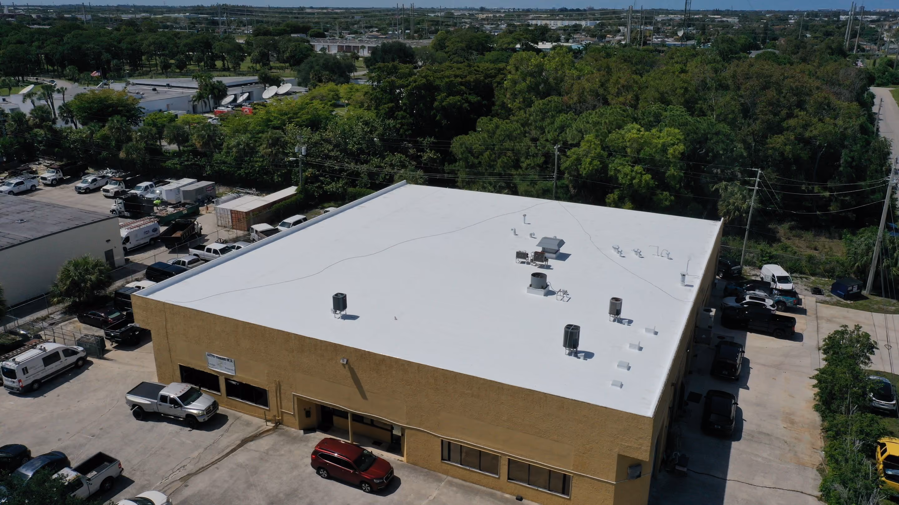 A white flat roof on a two-story tan commercial building seen from above, with rooftop units and vents, and vans and pickups parked around it