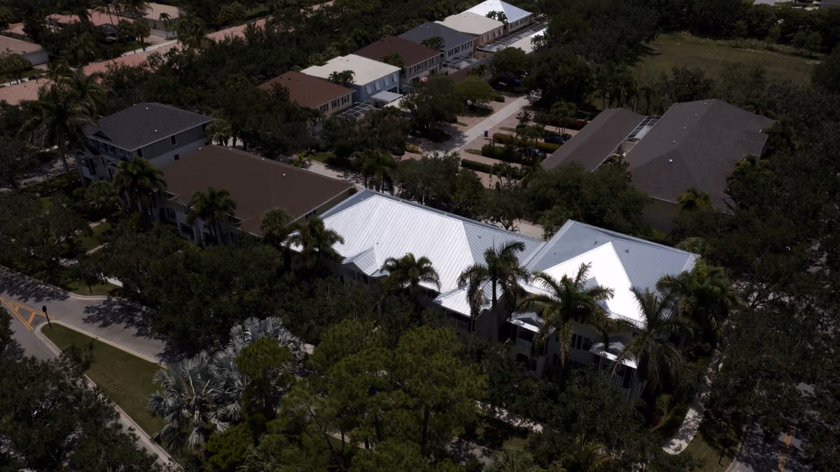 A long two-story building under a silver metal roof seen from the air among palms and trees, with shingle-roofed buildings behind it