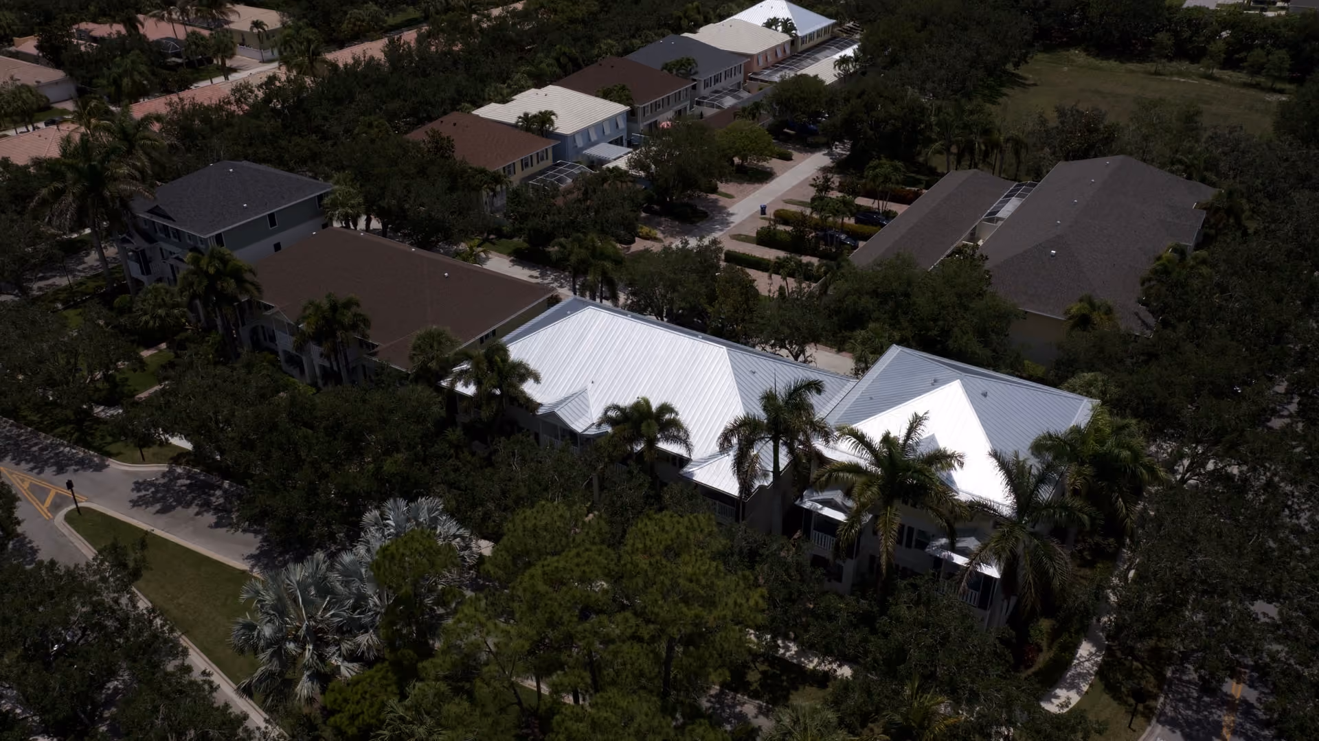 A condo building under a new white standing seam metal roof, seen from the air with mature palms and the community road below
