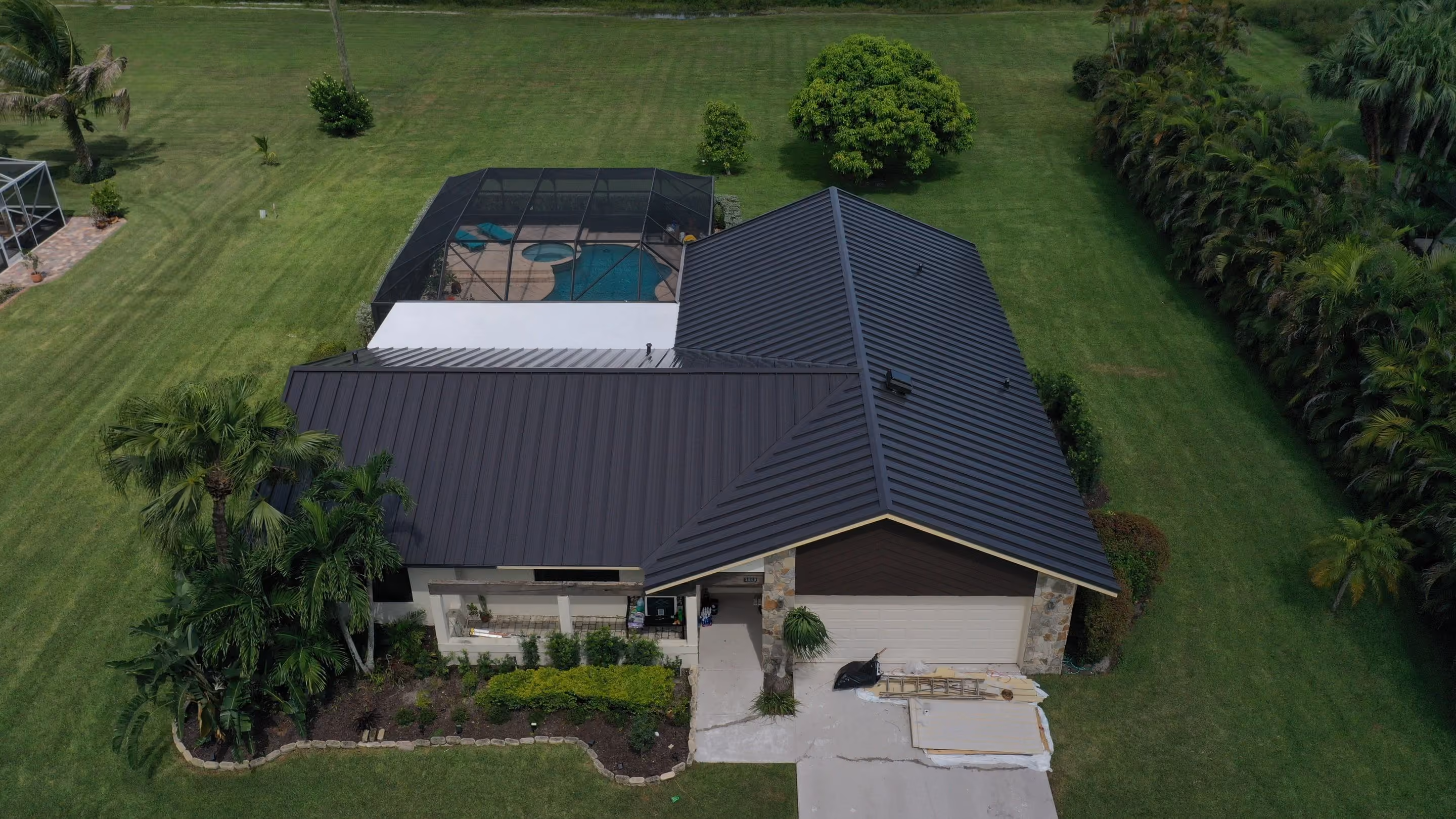 A single-story house with a black standing seam metal roof and a screened pool enclosure behind it, set on a wide mowed lawn